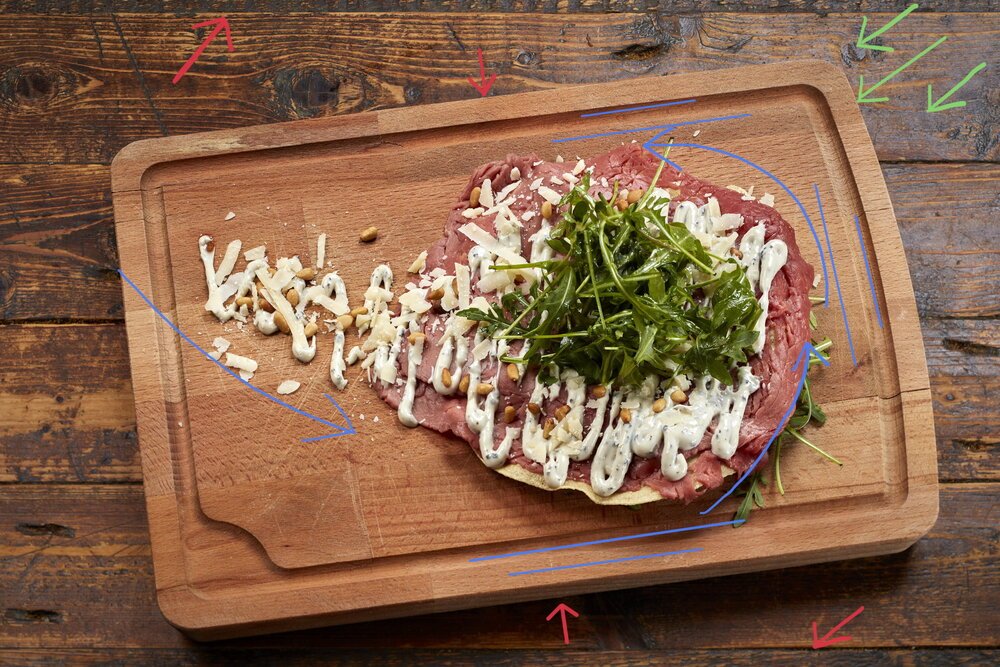 This photo has a bit more empty space. In composition theory this is called 'negative space'. Often this is used to create calm but it's also suitable for (marketing) texts and layout. The red lines show the background lines (deliberately horizontal). The board with carpaccio is slightly rotated for dynamism and promotion of the viewing direction (blue arrows.)